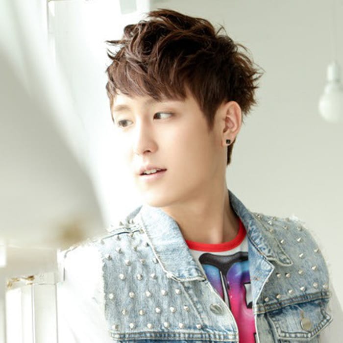 Iz has short brown hair, and he is wearing a light blue denim vest adorned with spikes, along with a white t-shirt underneath with blue-to-pink graphics or letters.