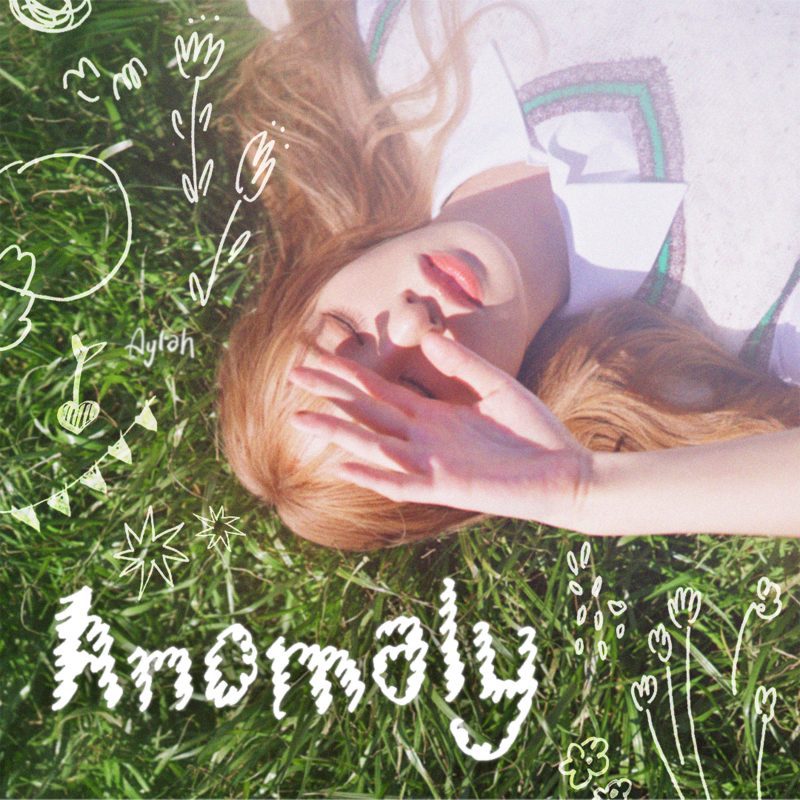 aylah laying on grass, covering her eyes from the sun with her hand. there is graphic text of the word 'anomaly' in the bottom left corner